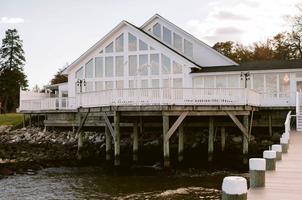 White waterfront house with large glass windows and wraparound deck built on stilts over rocky shoreline.