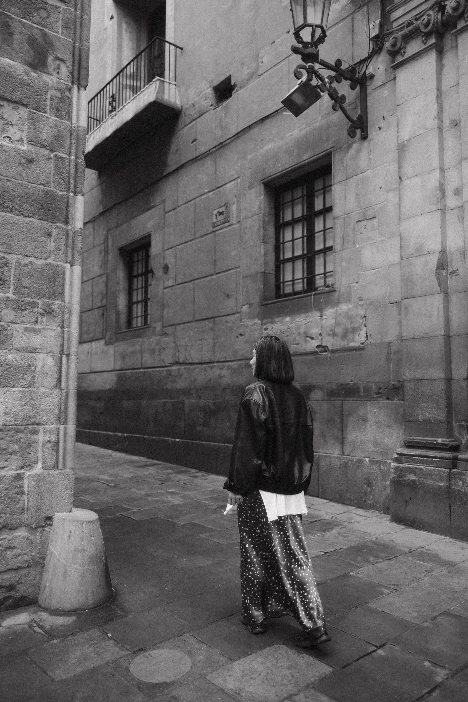 Black and white photo of a woman in floral pants and leather jacket standing in a historic stone alleyway.