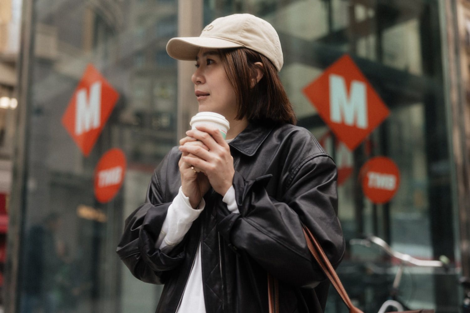 Young woman in black jacket and cap holding coffee cup near metro station entrance.