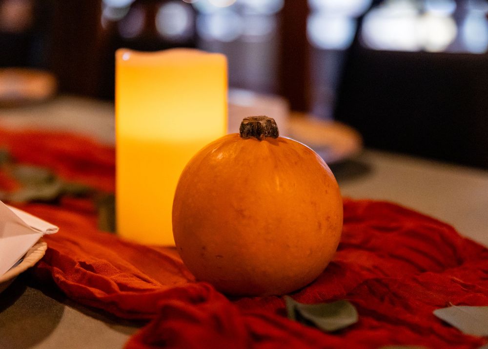 Small orange pumpkin beside a glowing candle on a red table runner, autumn fall wedding centerpiece decor.