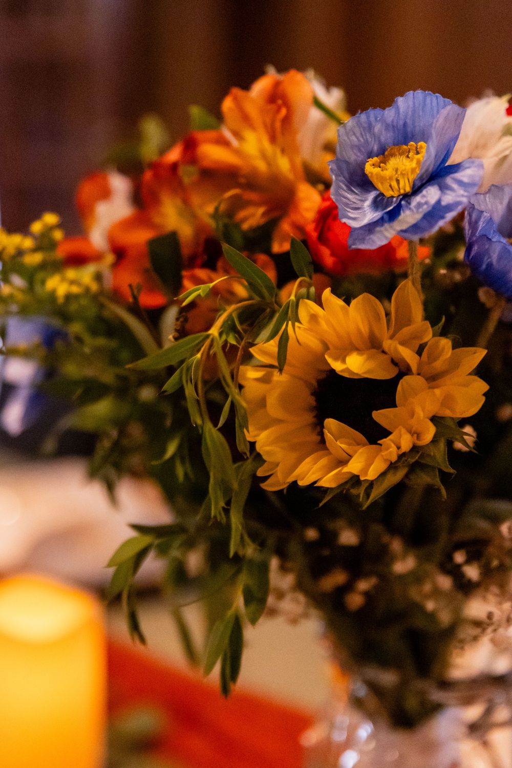 Colorful wedding centerpiece bouquet featuring blue poppies, orange alstroemeria, and yellow sunflowers with a candle nearby.
