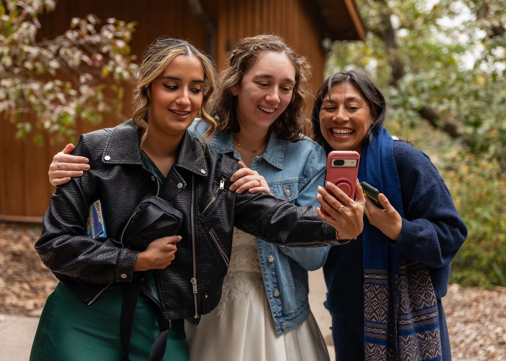 Three women smiling and taking a selfie outdoors, two in leather jackets and one in a blue shawl at Platte River State Decker Creek Lodge
