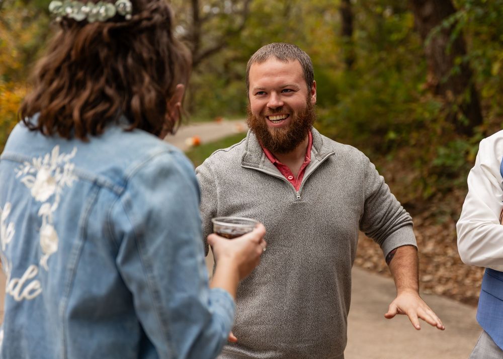Bearded man in gray pullover smiling warmly during an outdoor autumn gathering with guests.