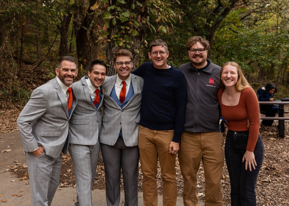 Group of six people smiling together on a wooded path during autumn, dressed in suits and casual attire.