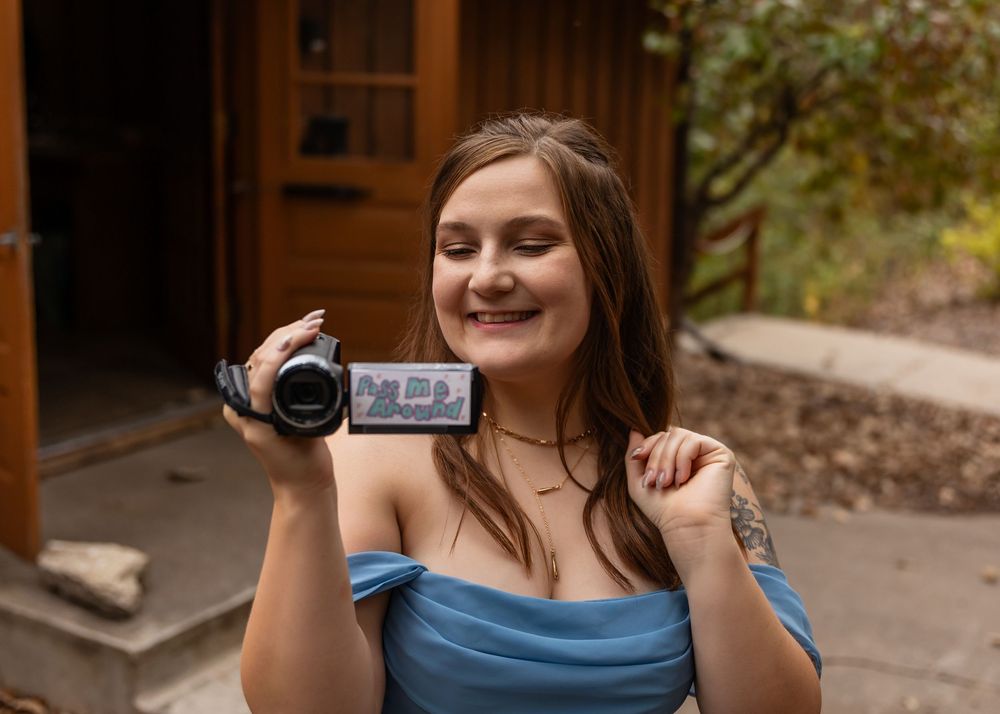 Young woman in blue off-shoulder dress smiling while holding a Sony camcorder outdoors near Decker Creek Lodge at Platte River State Park