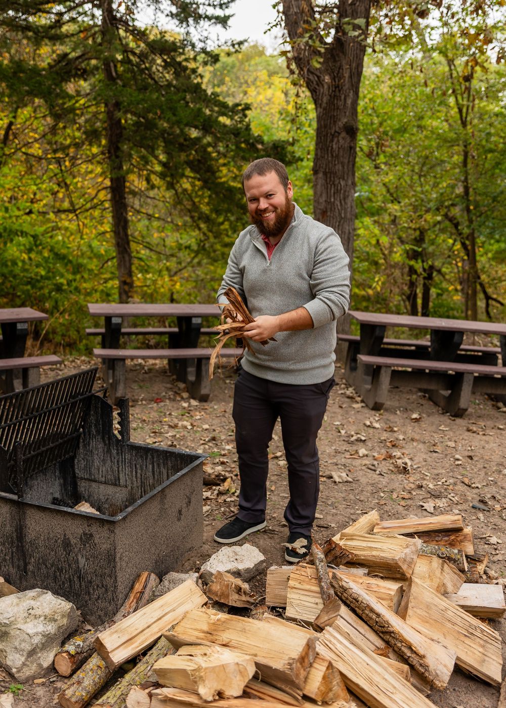 Bearded man smiling while splitting firewood at an outdoor campfire pit surrounded by autumn trees.