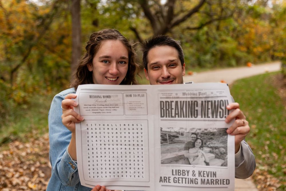 Smiling couple holds a wedding newspaper announcing Libby and Kevin are getting married outdoors in fall.
