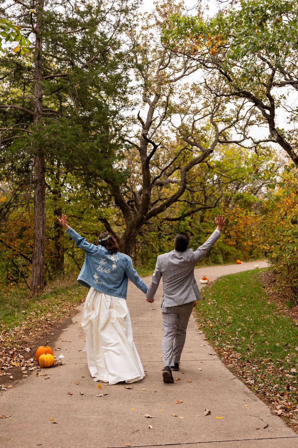 Newlywed couple walking hand-in-hand down autumn path, raising hands joyfully, bride in white gown and denim jacket.