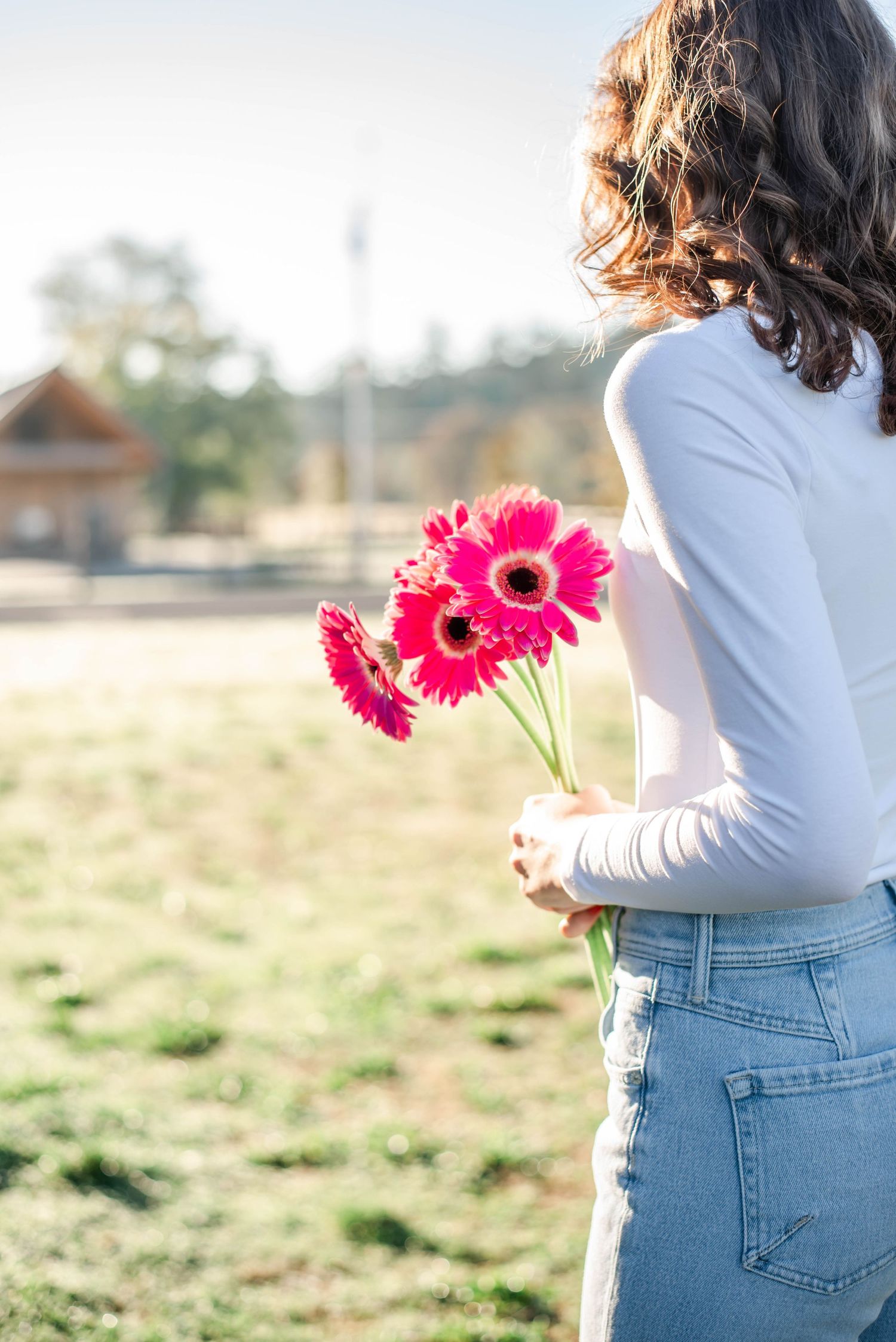 Woman in white top and jeans holding vibrant pink gerbera daisies behind her back outdoors.