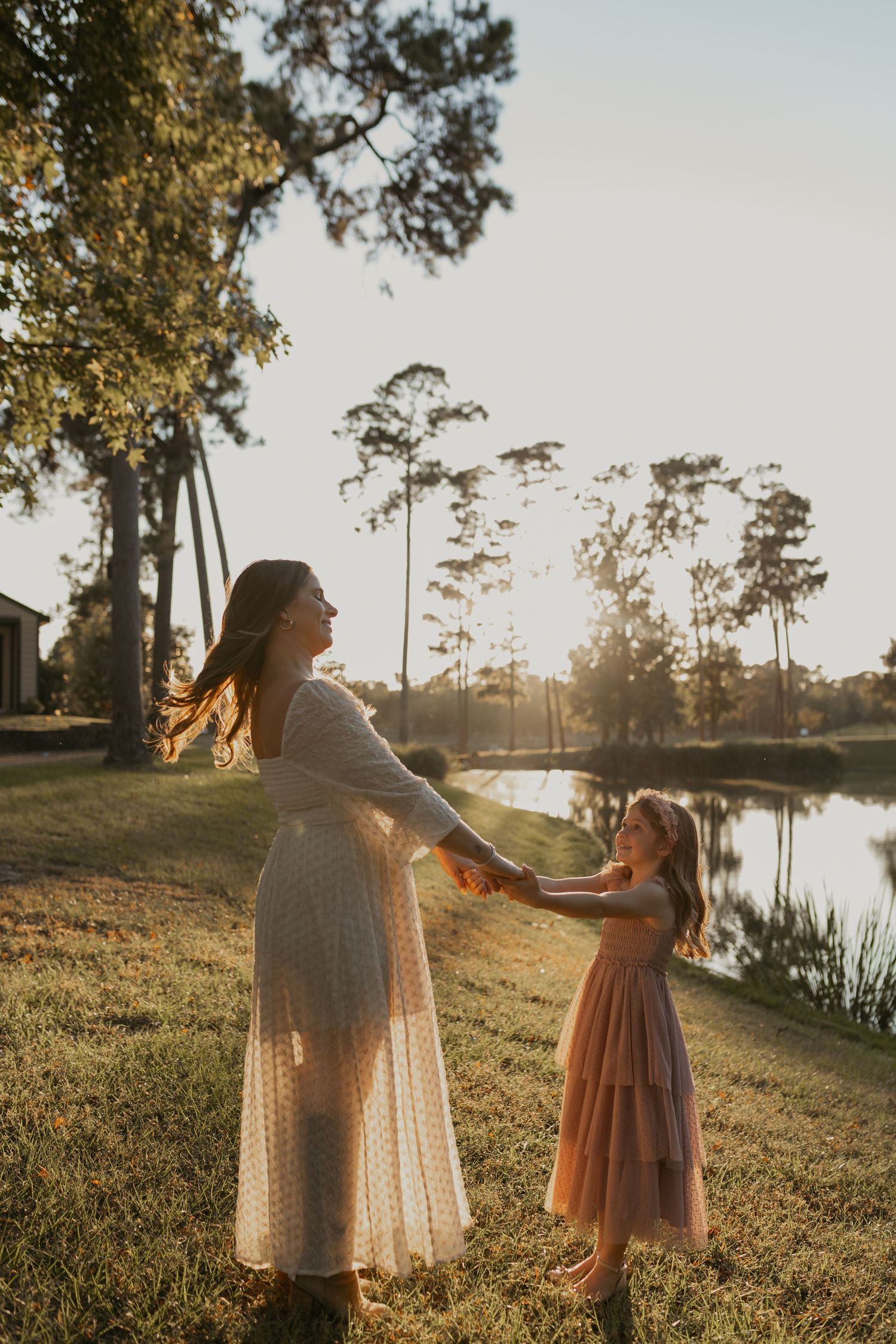 A tender moment captured at sunset by a lake as two figures in flowing dresses dance together on the grassy shore.