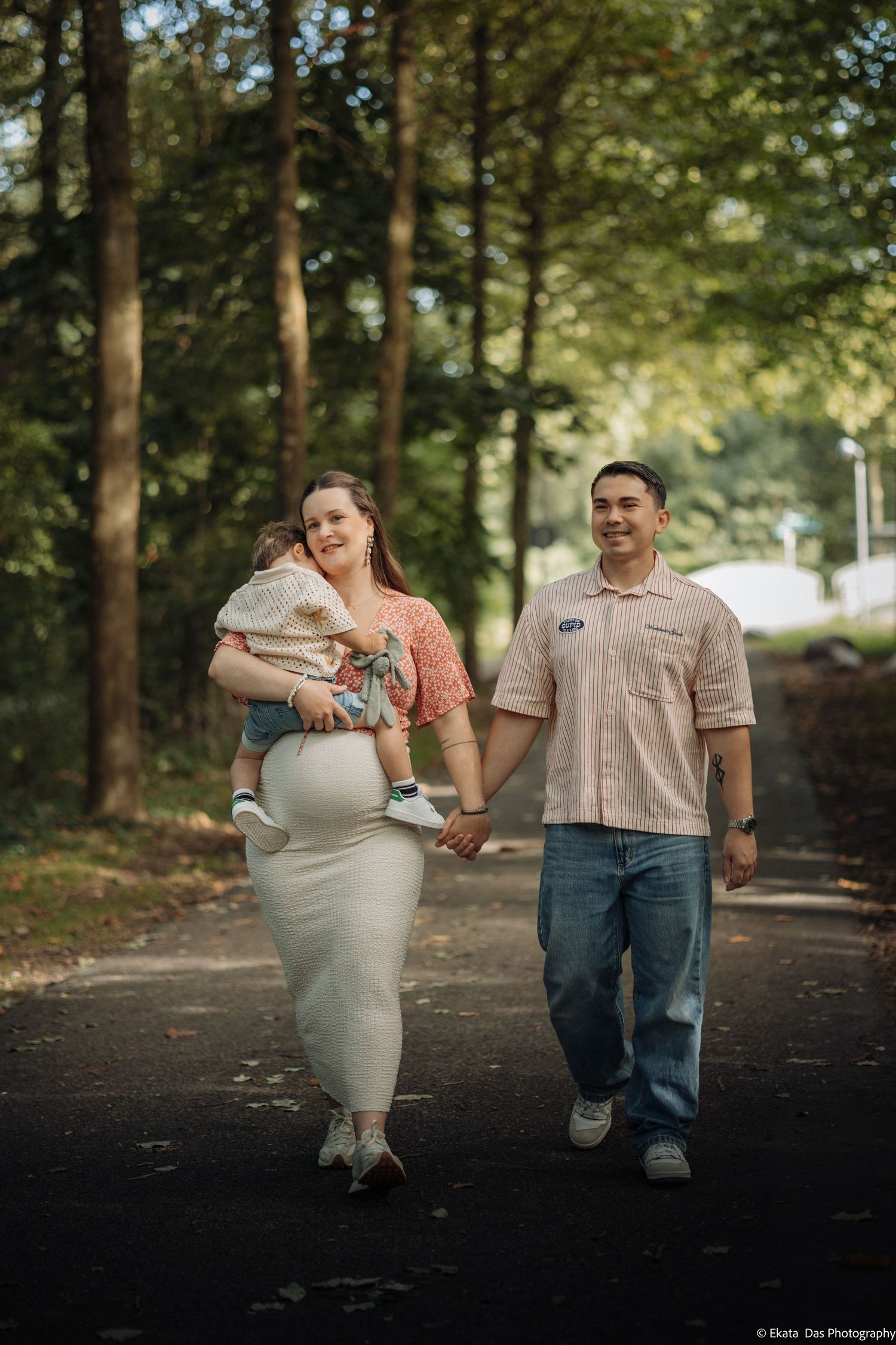 A smiling couple holds hands and walks along a tree-lined path, the pregnant woman carrying a toddler in her arms.