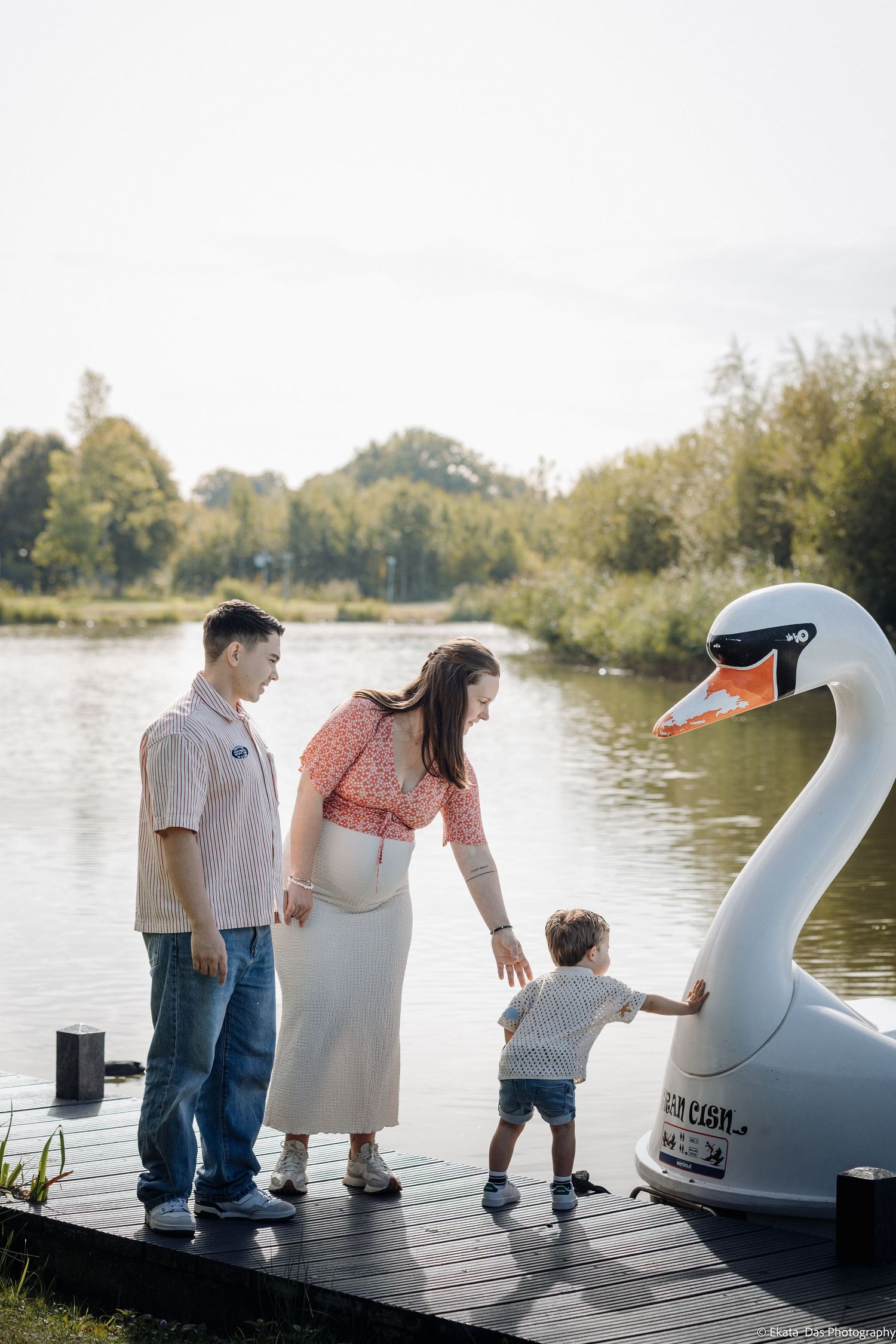 A family of three stands on a lakeside dock as a toddler reaches out to touch a white swan pedal boat on a sunny day.