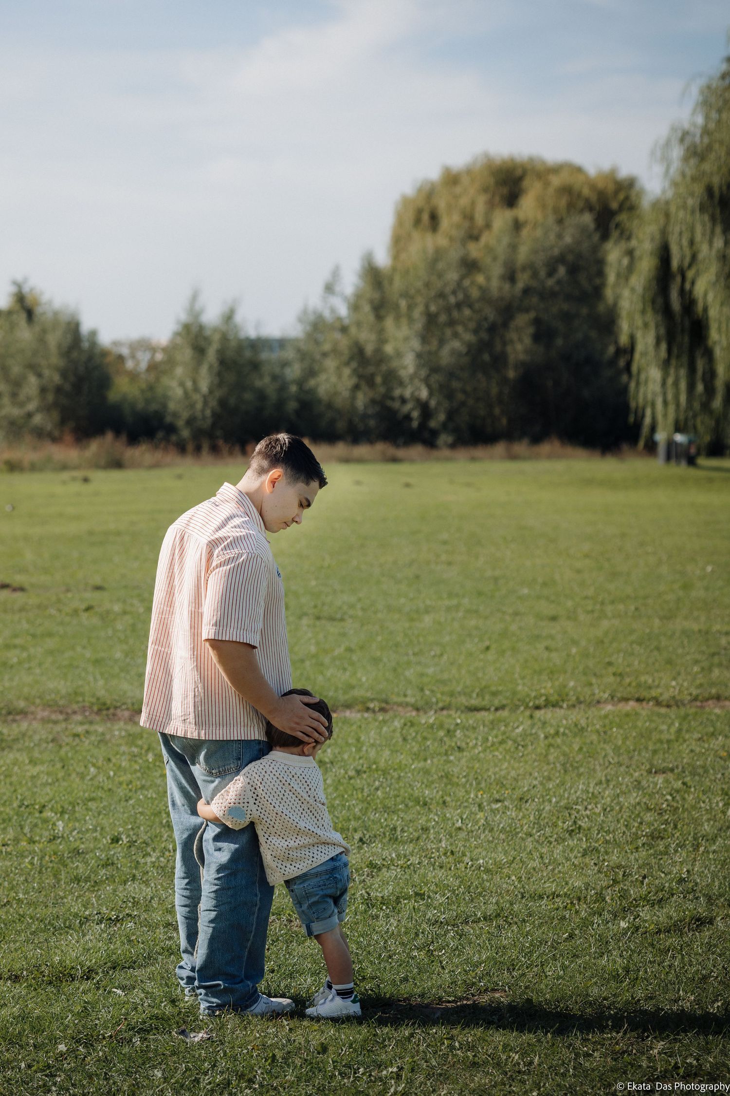 A man in a pink shirt and jeans stands on a grassy field with his hand on the head of a toddler hugging his leg.
