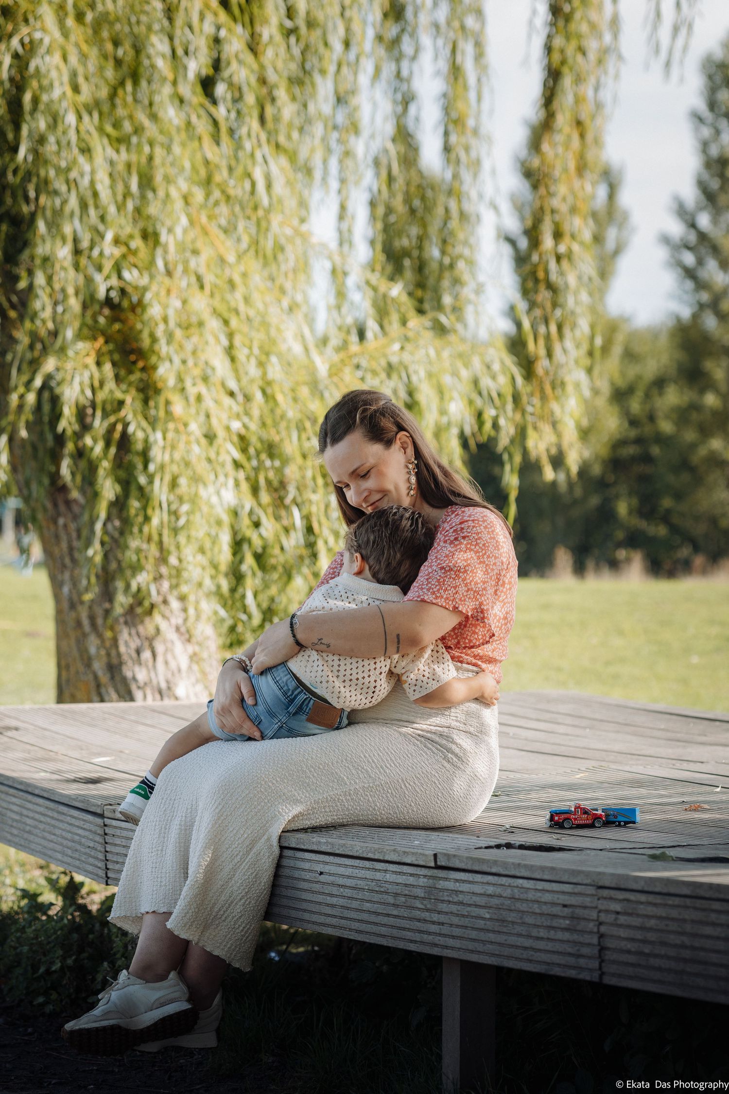 A woman in a coral top and cream pants sits on a wooden platform under a willow tree, hugging a young child close.
