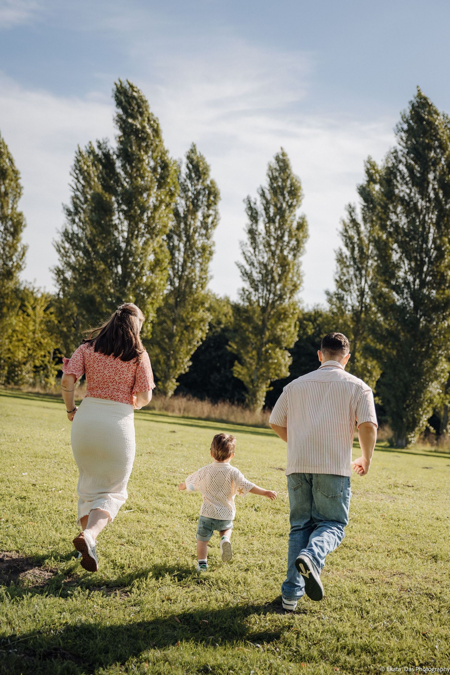Family of three running together across a sunny grassy park with tall poplar trees, a toddler between two parents.
