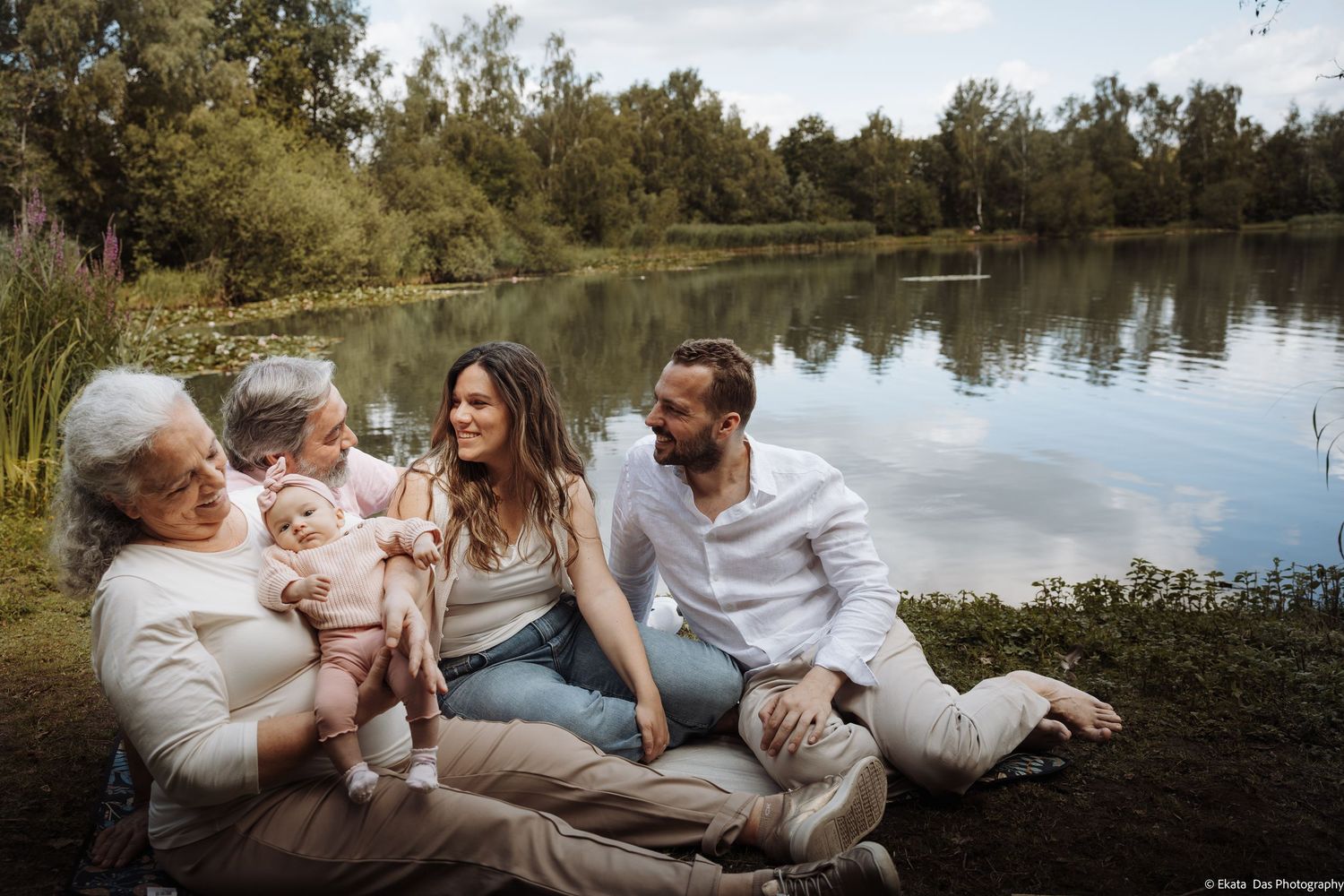 Multi-generational family sitting by a serene lake, sharing a joyful moment with a baby outdoors.