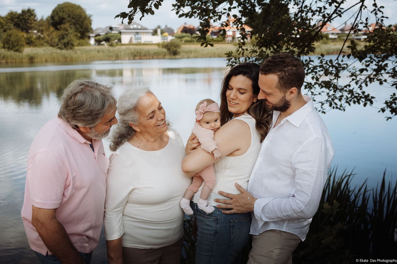 Multi-generational family portrait outdoors by a lake, featuring grandparents, parents, and a baby in a serene setting.