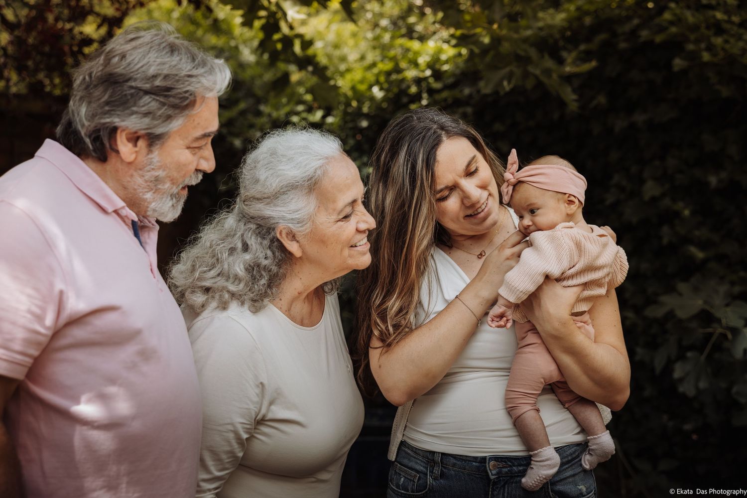 Multigenerational family smiling and admiring a newborn baby outdoors, surrounded by lush green foliage.