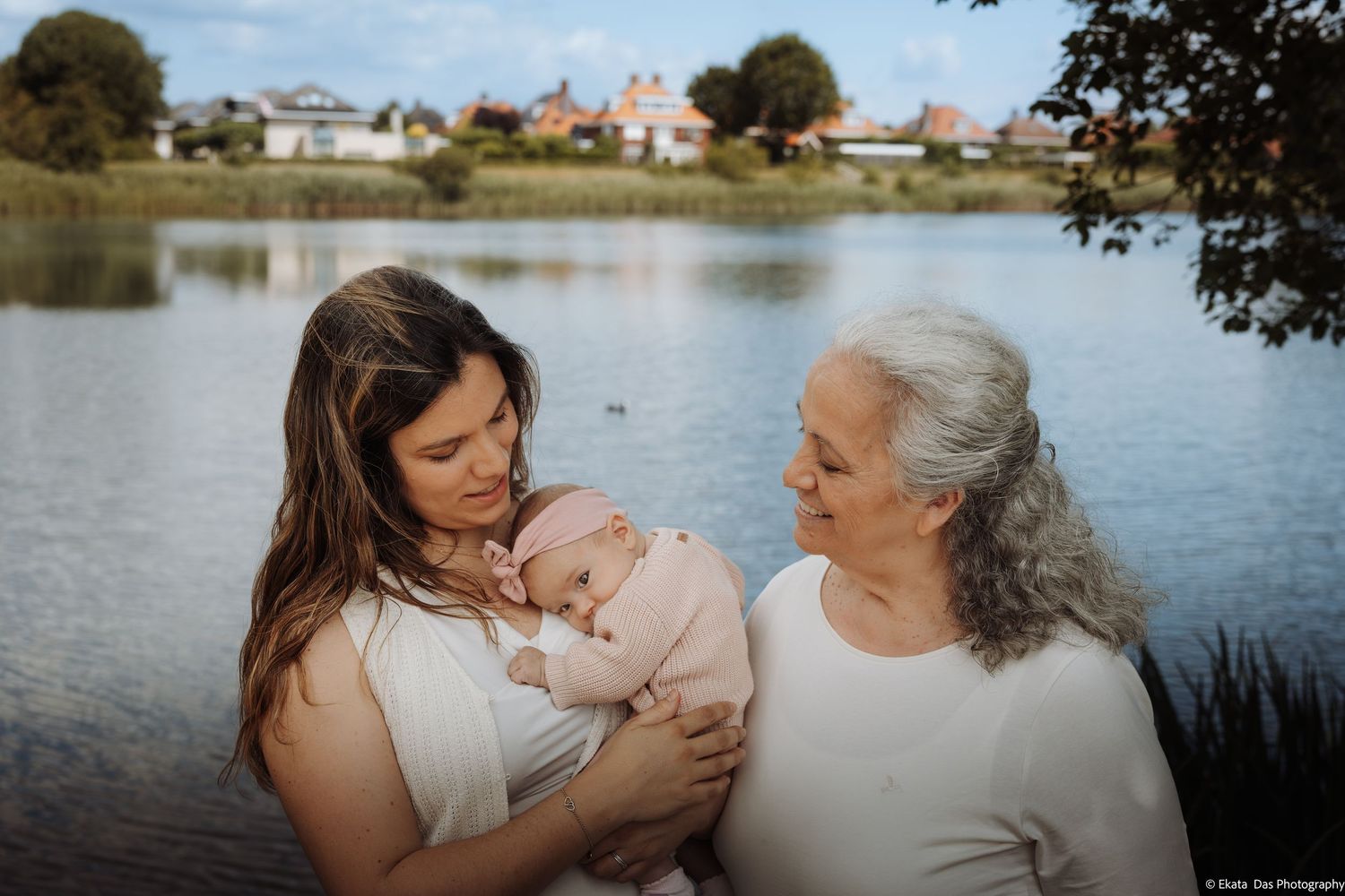 A mother holds her newborn baby while an older woman looks on lovingly near a serene lake.