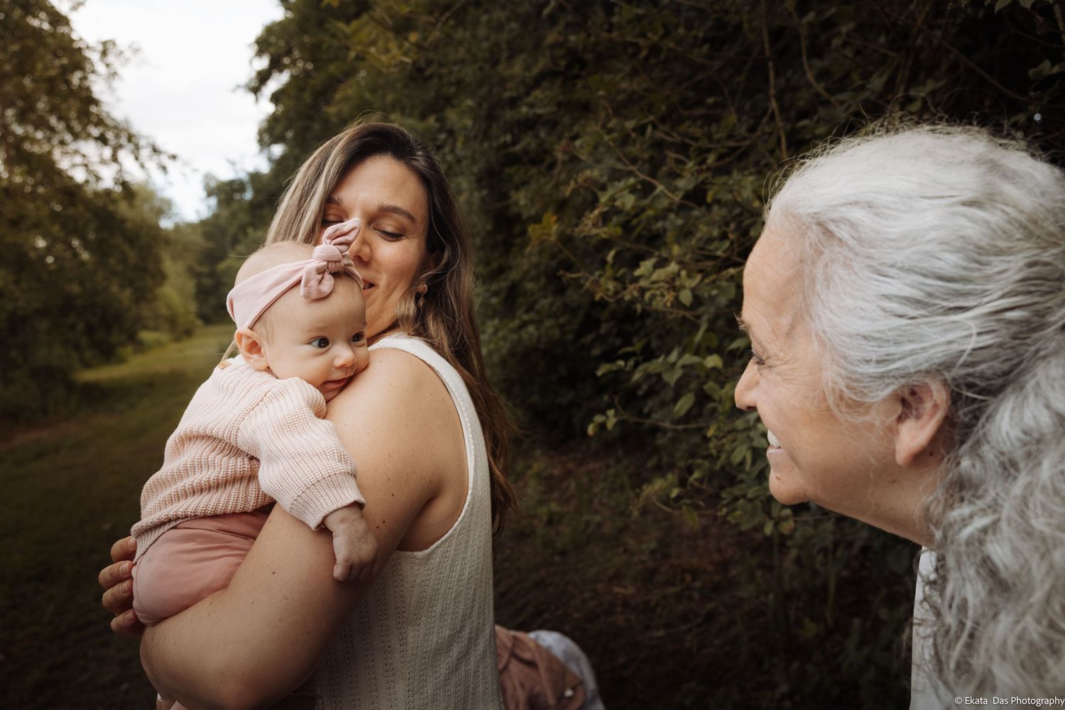 A mother holds her baby girl while an older woman smiles at the infant outdoors in a lush green setting.