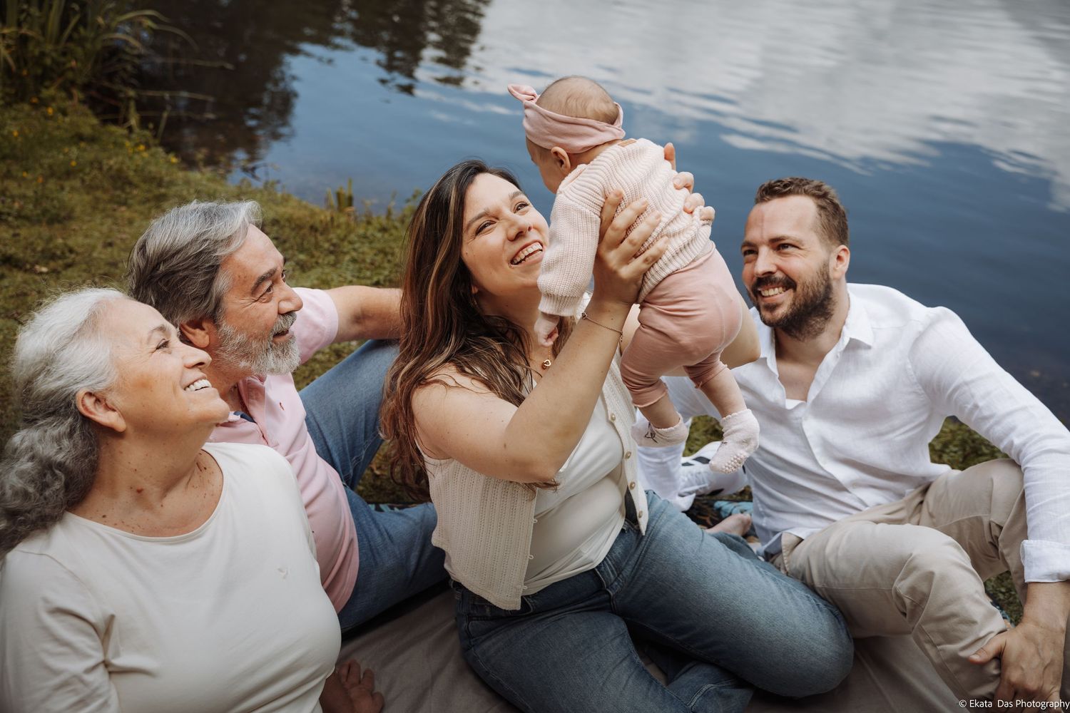 Multi-generational family laughing together outdoors by a lake, with parents lifting a smiling baby girl.