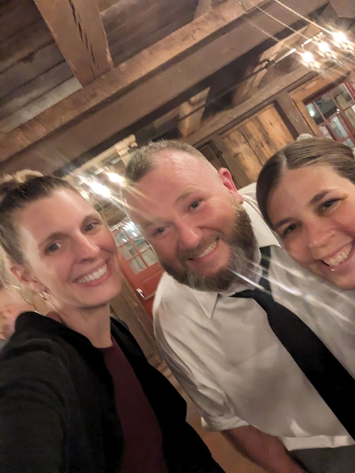 Three friends smiling together for a selfie in what appears to be a rustic indoor setting with wooden beams visible above.