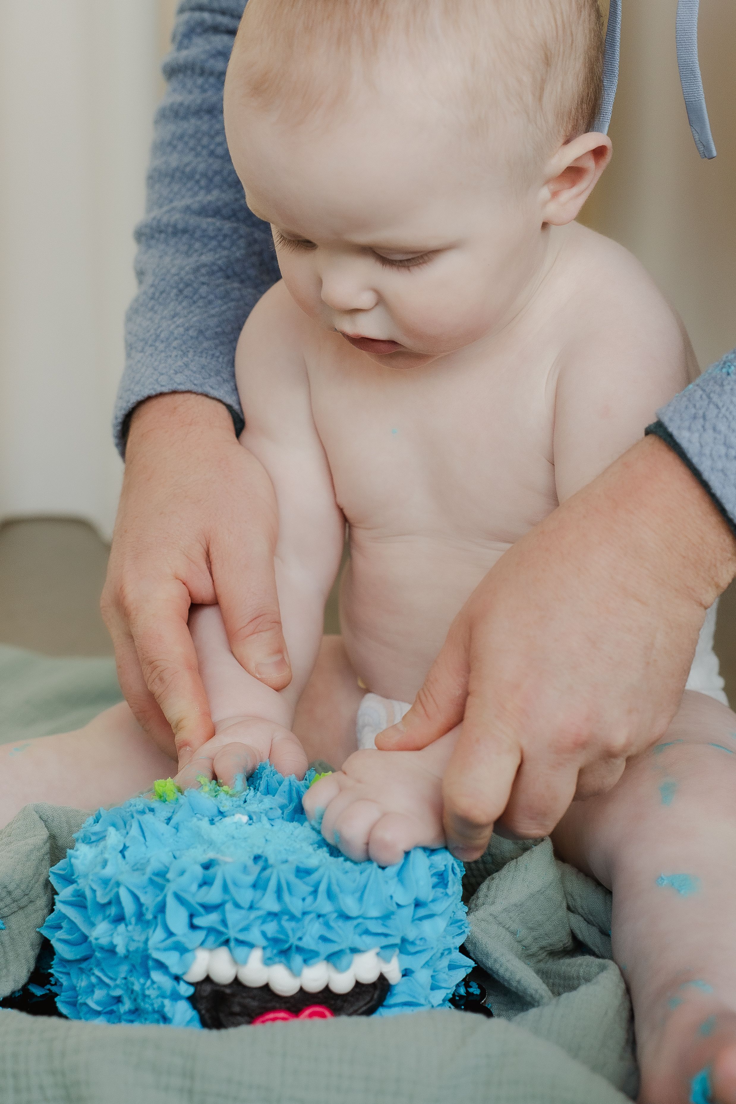Baby's hands reach for bright blue frosted smash cake during first birthday photoshoot.