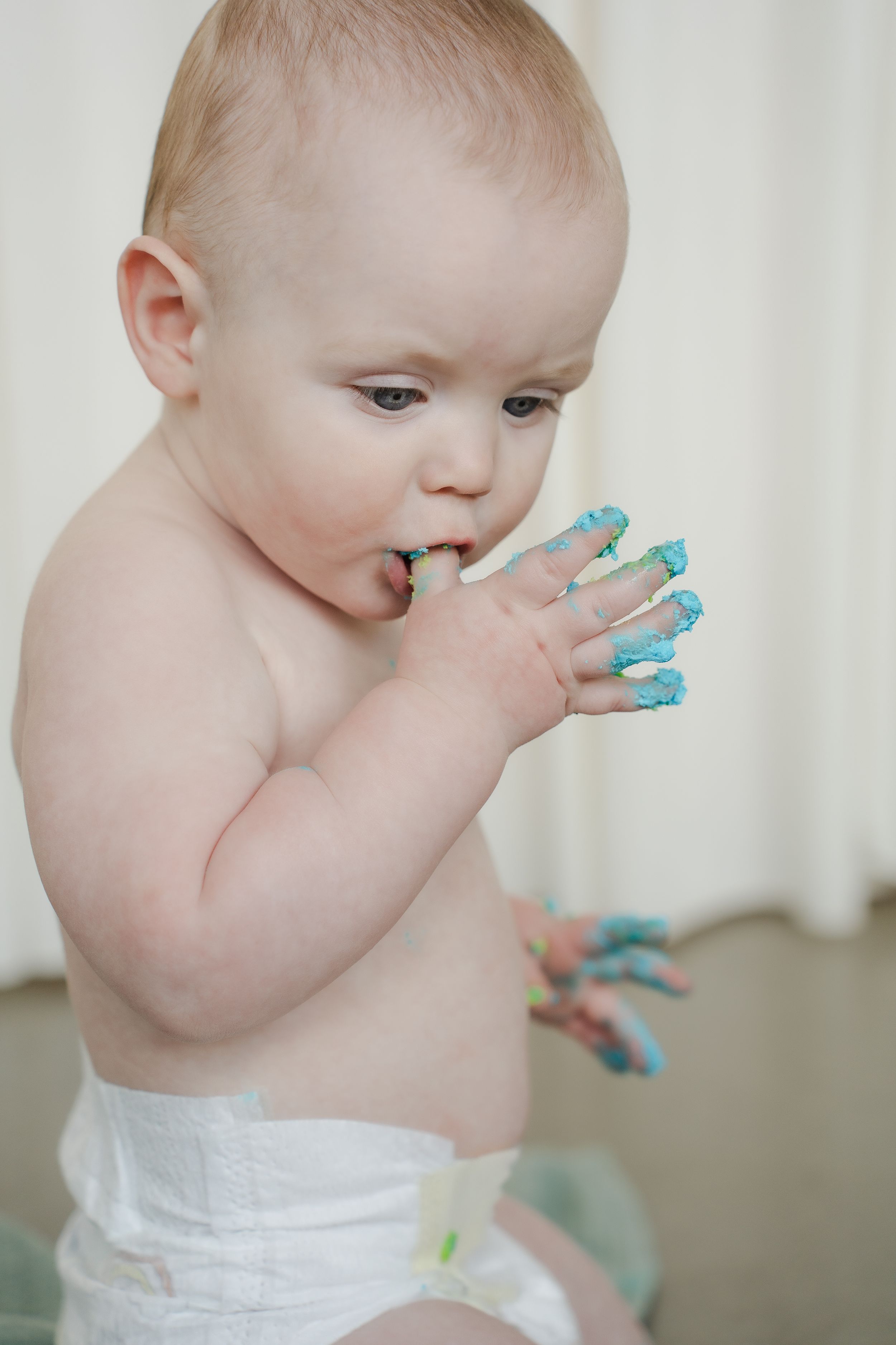 Baby in white diaper chews on blue teething toy while standing.