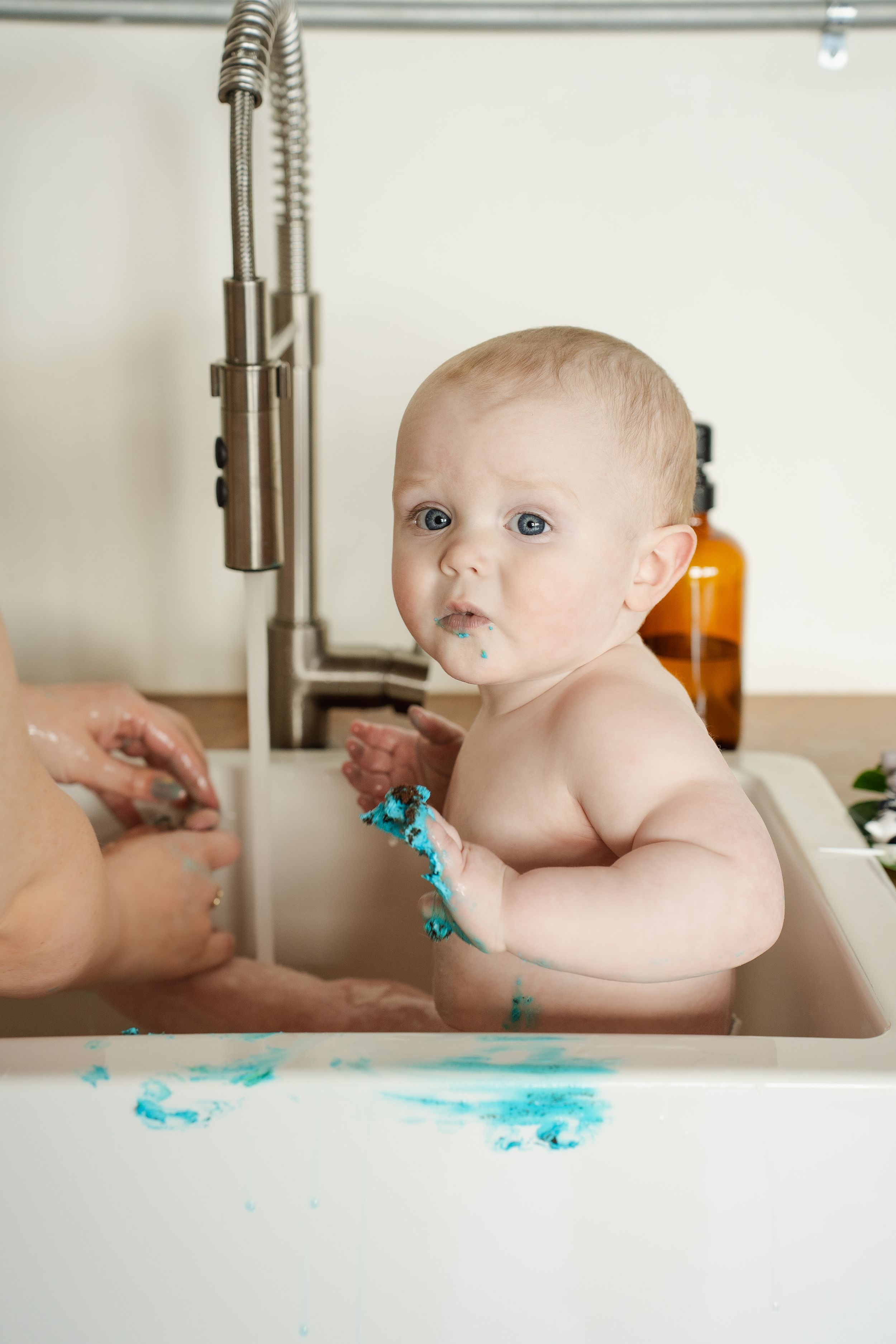 Baby in a kitchen sink plays with blue frosting or paint, creating a messy but fun sensory experience.