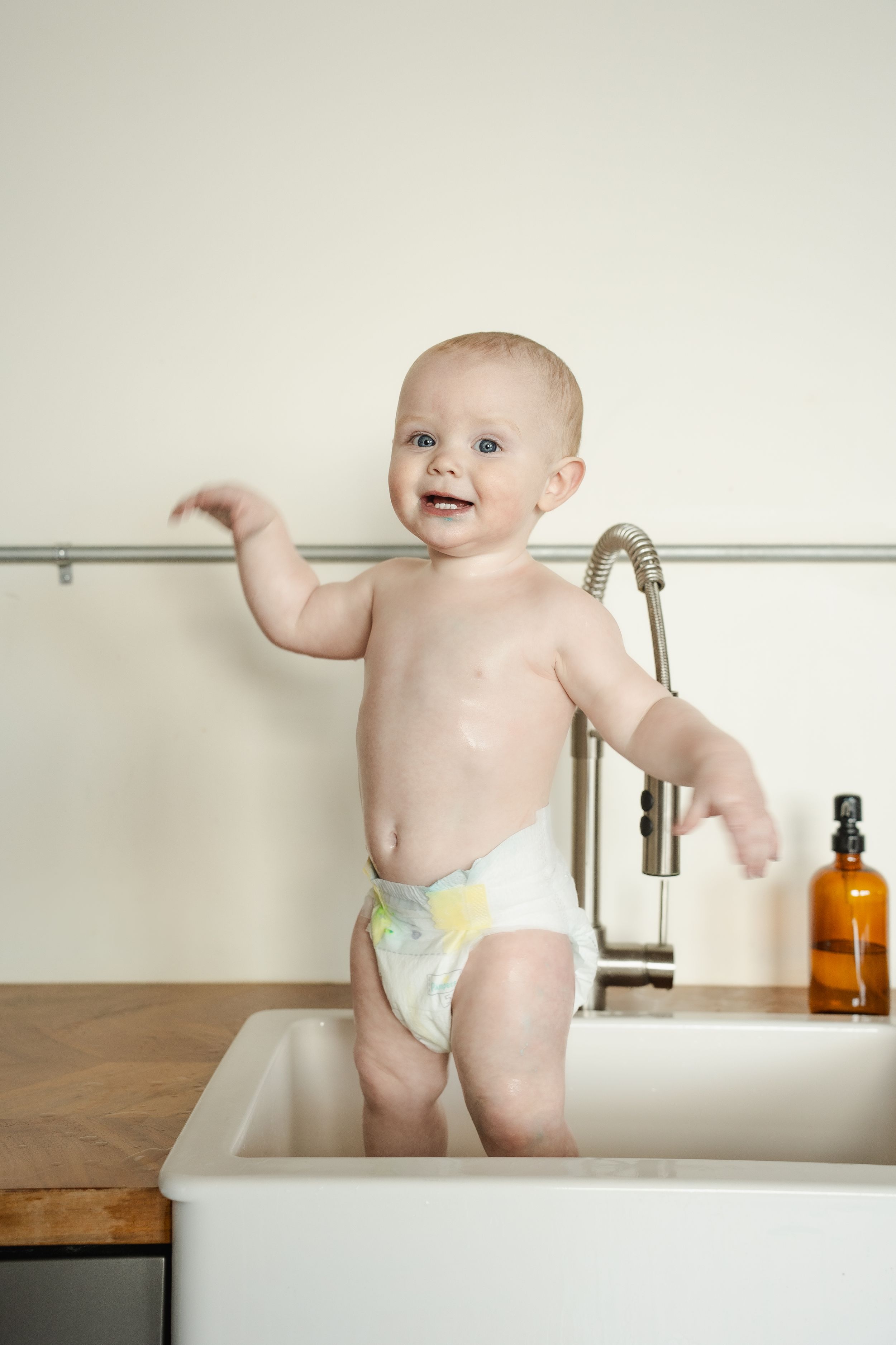 Baby in diaper plays and smiles while standing in white kitchen sink with amber bottle in background.