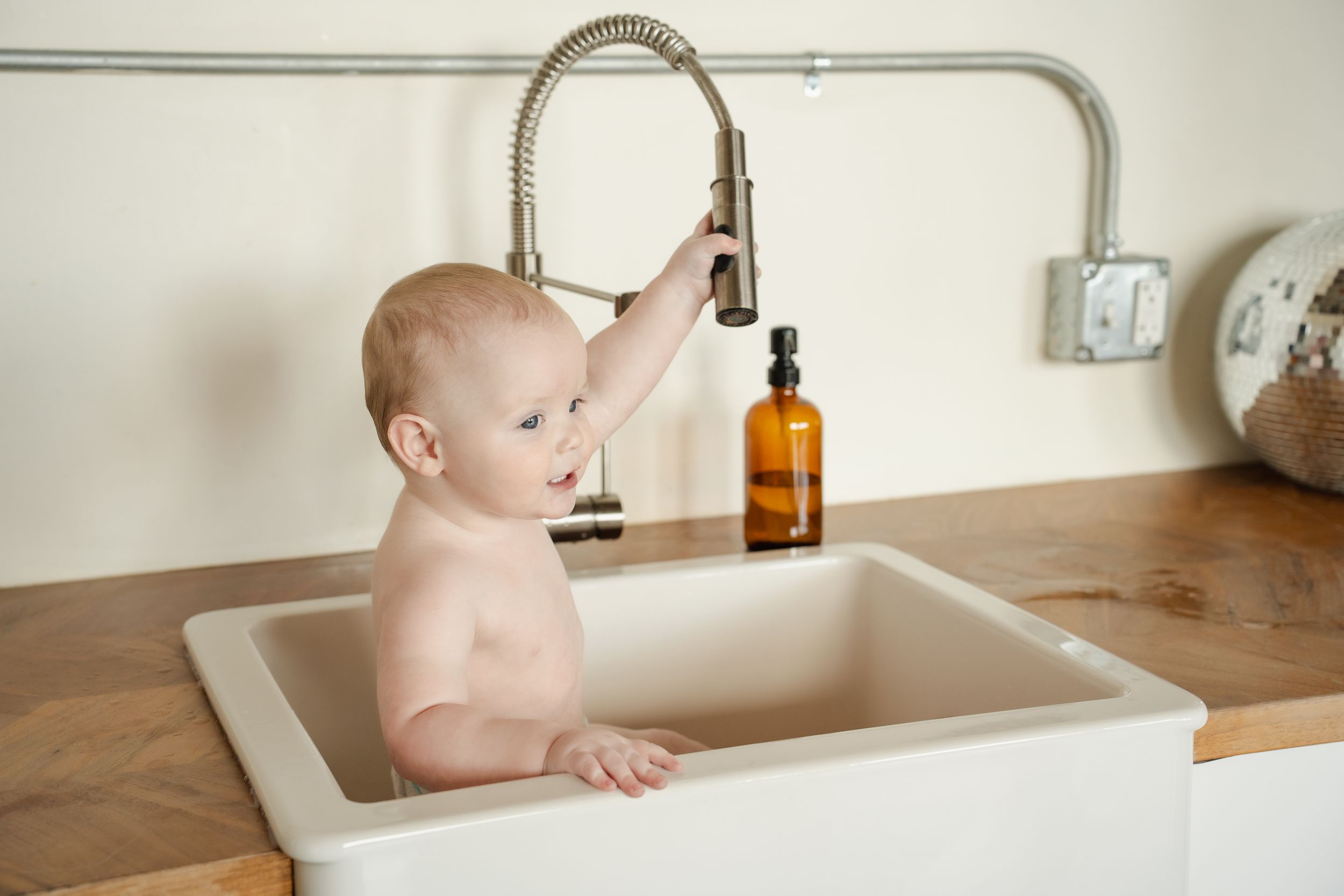 Baby stands in farmhouse sink with modern faucet and wooden countertop.
