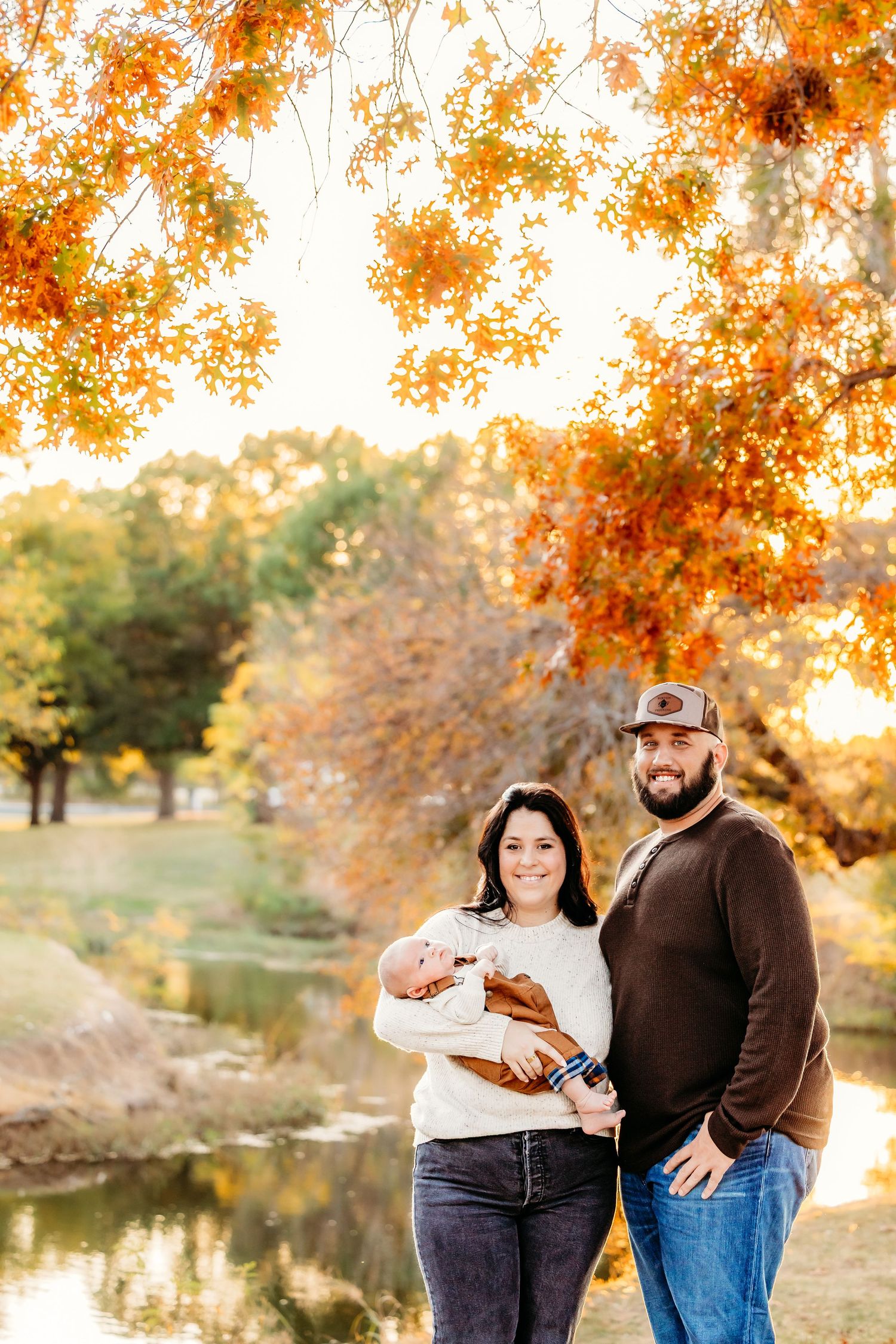 Couple stands together holding their infant outdoors during autumn with orange fall foliage and a creek in the background in Keller, TX.