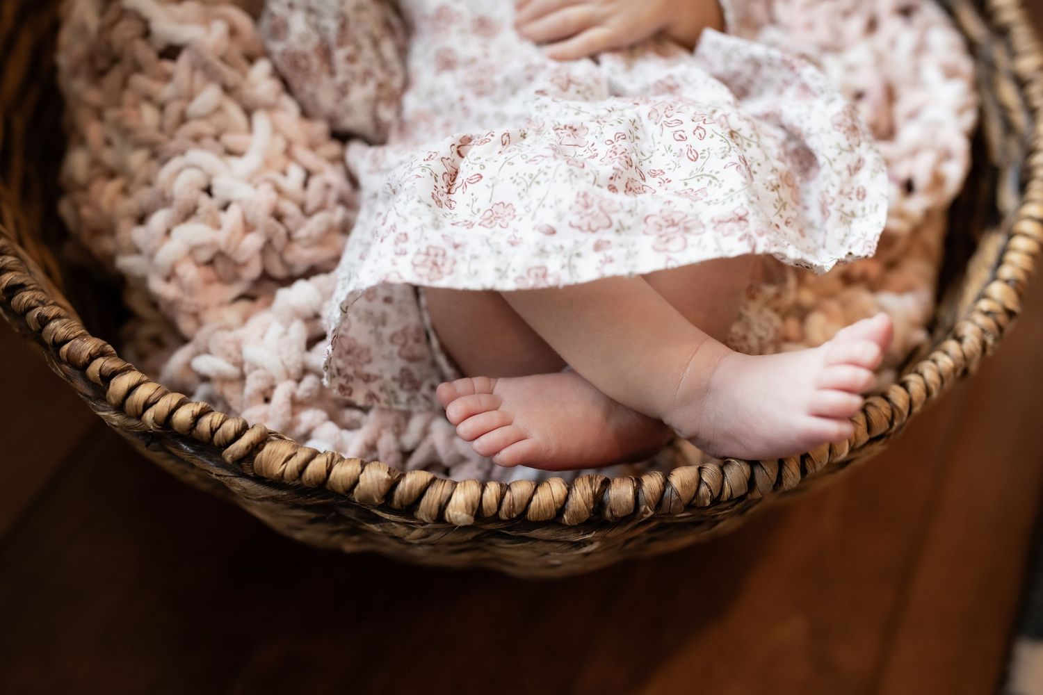 Newborn baby bare feet peeking from floral dress in a wicker basket with pink blanket.