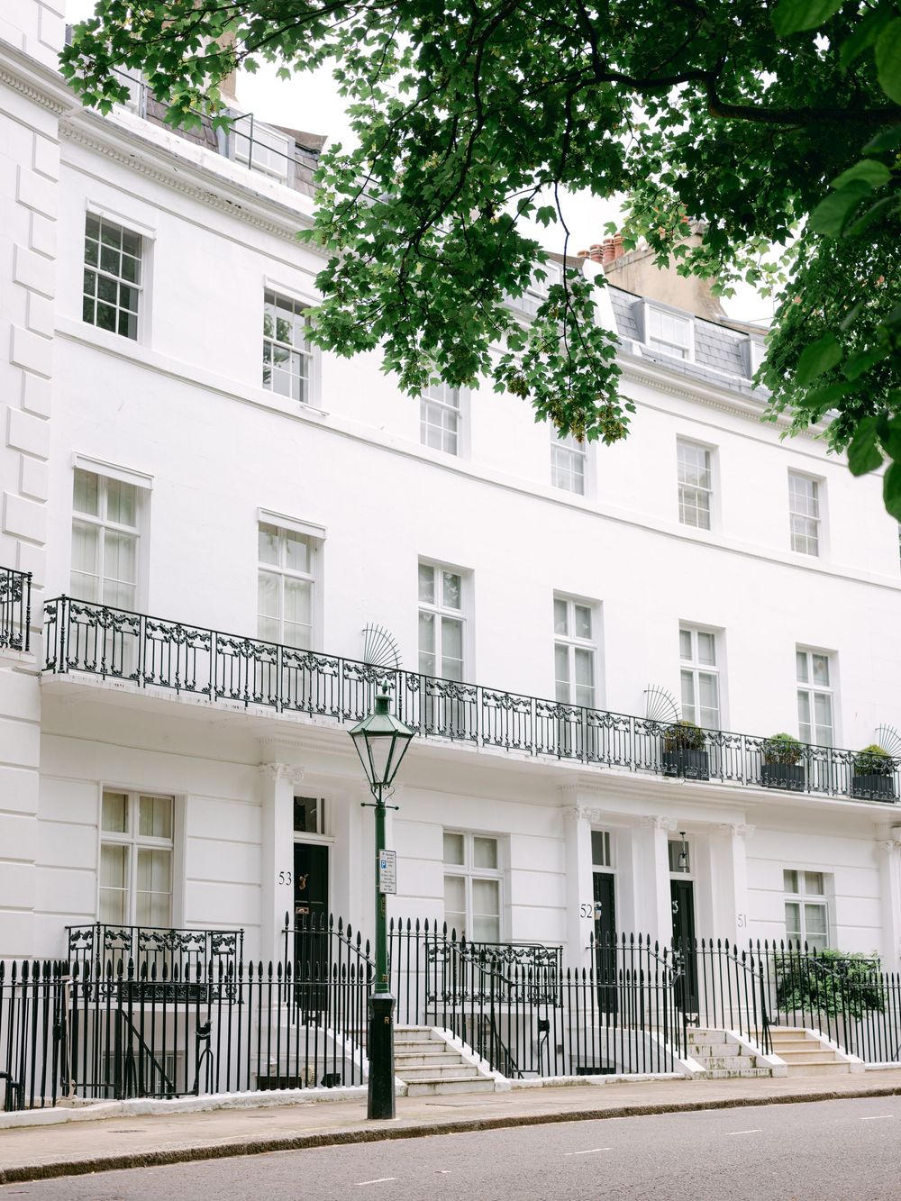White Georgian townhouse with black iron railings and balconies, street lamp, and tree-lined pavement in London.