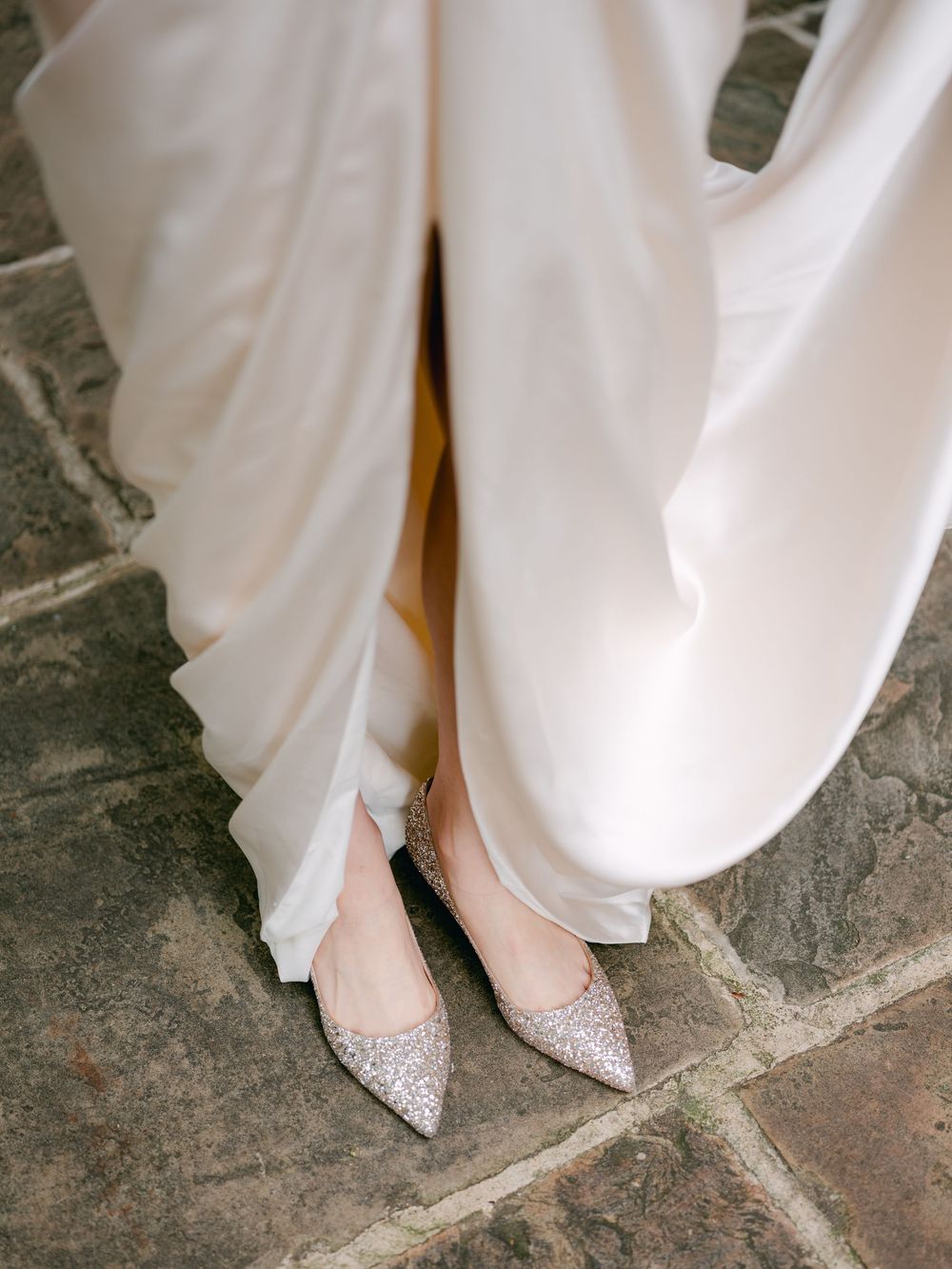 Bride wearing sparkling silver glitter pointed-toe flats peeking beneath a flowing champagne satin wedding gown on stone pavement.
