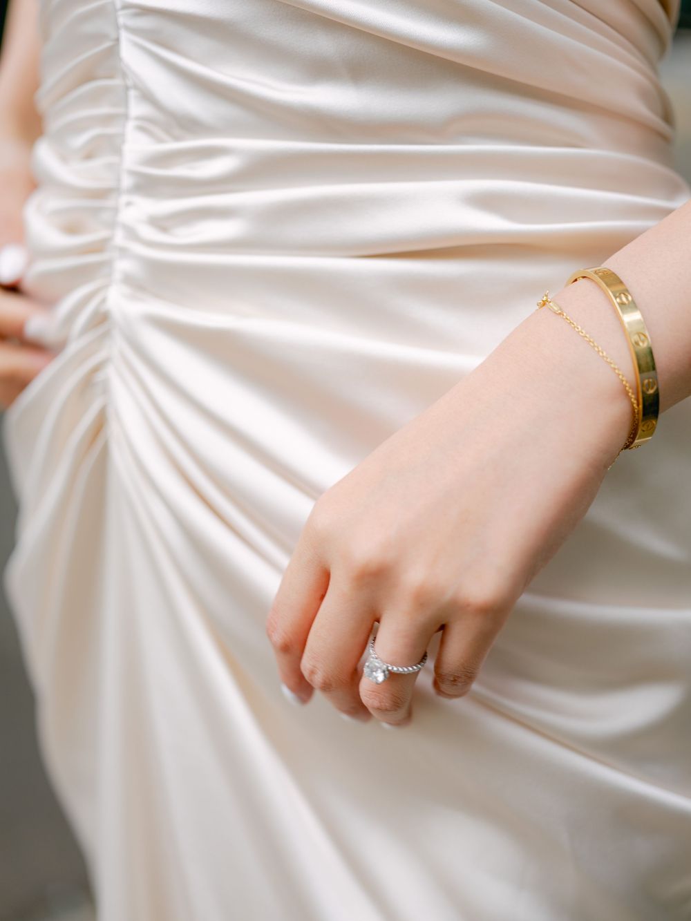 Close-up of bride's hand showing diamond engagement ring and gold Cartier bracelet on satin wedding dress.