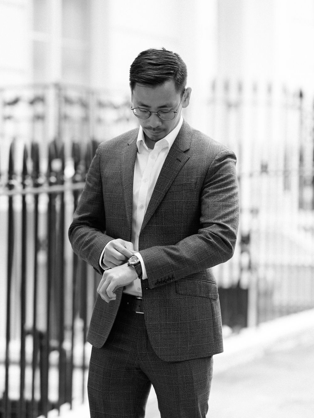 Black and white photo of a man in a suit checking his watch outdoors near iron railings.