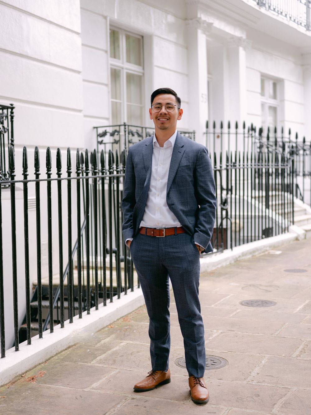 Man in navy blue suit with brown leather belt and shoes stands confidently on a London street beside wrought iron fence.