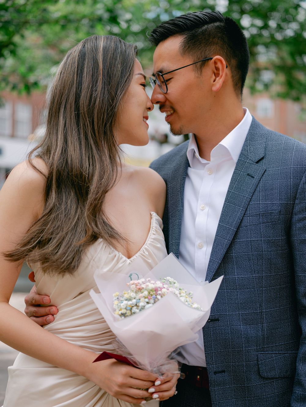 Couple in wedding attire share a romantic moment, woman in white dress holds floral bouquet, man in gray suit outdoors.