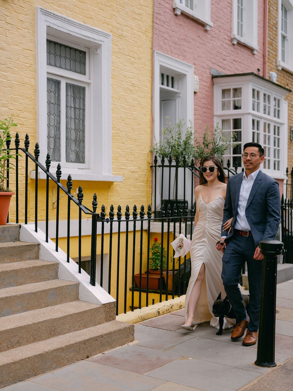 A couple in elegant attire poses on colorful Notting Hill street steps, woman in champagne slit gown, man in blue suit.