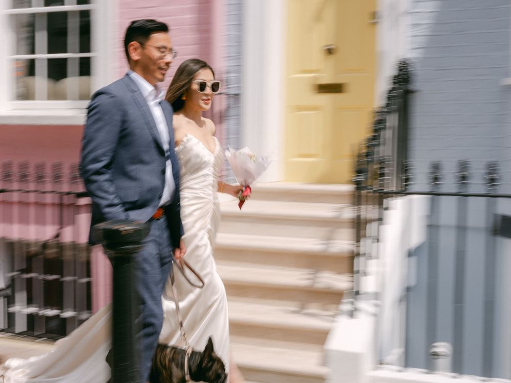 Laughing couple walking past pastel-colored townhouses in Notting Hill, bride in satin dress with sunglasses.