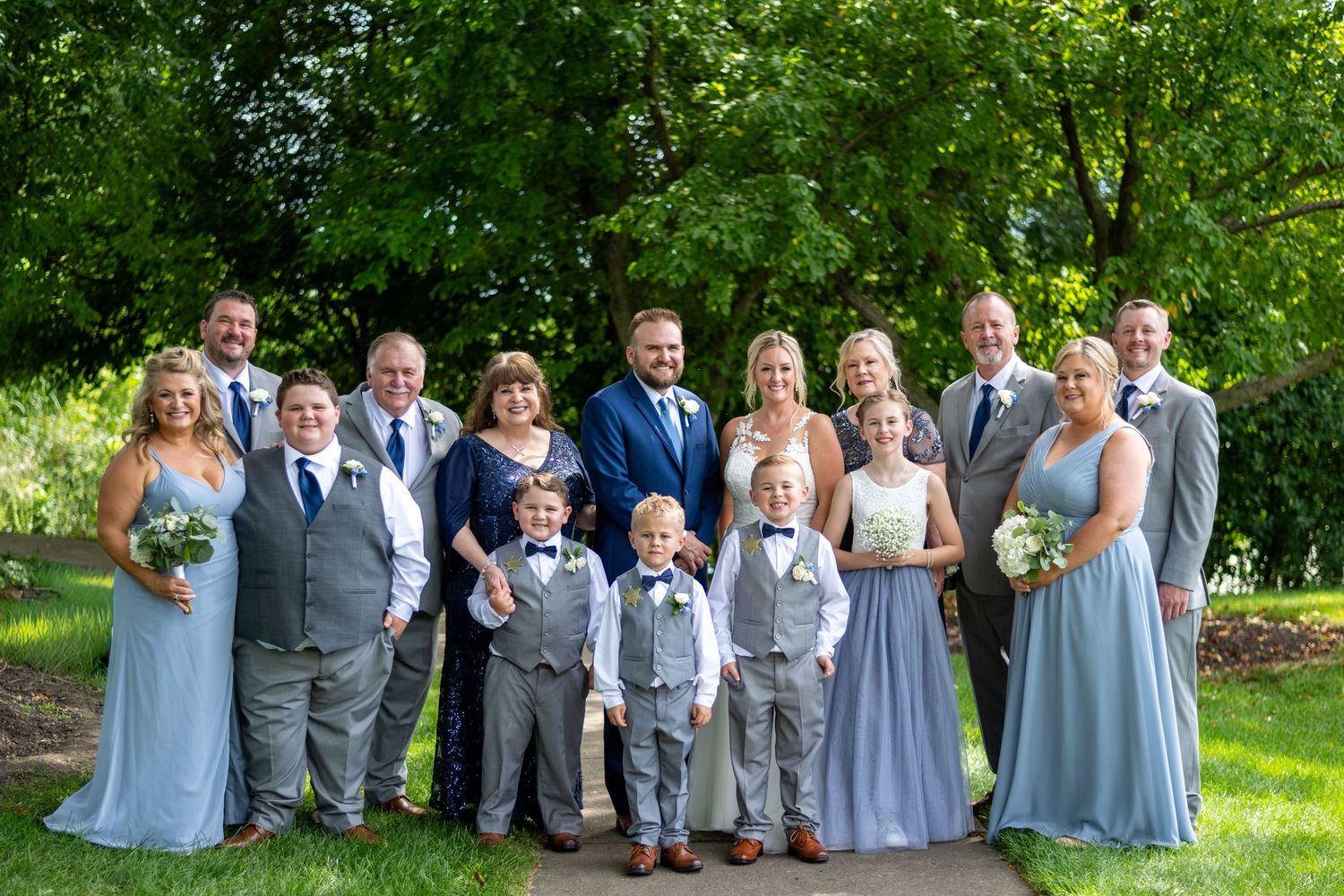 Wedding party in light blue dresses and gray suits poses together on a lush green lawn during a summer celebration.