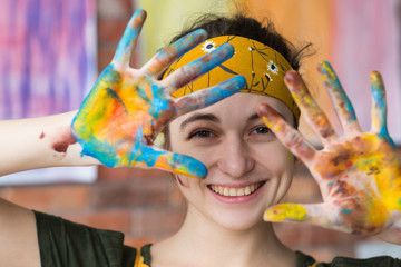 Smiling young woman with paint-covered hands raised showing colorful palms, wearing yellow headband in art studio.