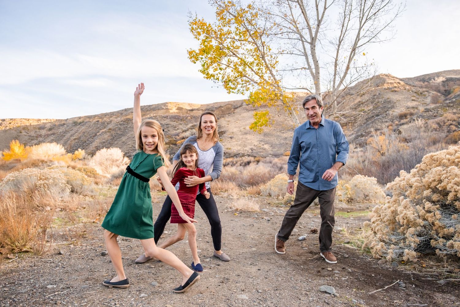 Family of four posing playfully on a desert hiking trail surrounded by autumn foliage and rocky hills.