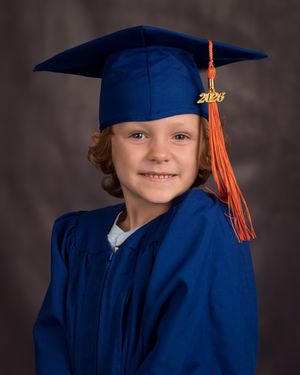 Young child in blue graduation cap and gown with orange tassel and 2023 charm smiles for portrait photo.
