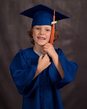 Young child smiling in blue graduation cap and gown with orange tassel, celebrating preschool graduation.
