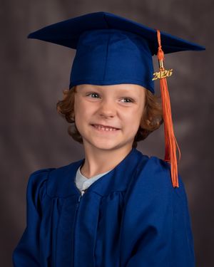 Young child in blue graduation cap and gown with orange tassel smiling for a 2026 graduation portrait photo.