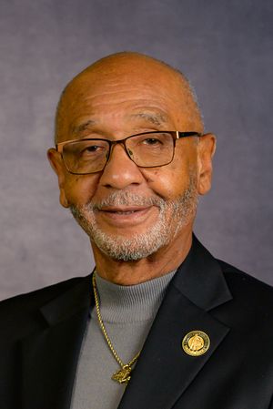 Elderly man with glasses and gray beard wearing a black blazer with gold pin and chain necklace, formal portrait.