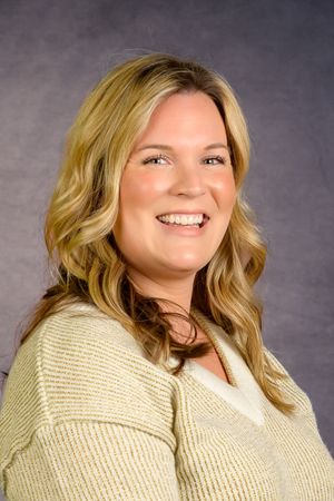 Smiling blonde woman in a cream knit sweater poses for a professional headshot against a gray background.