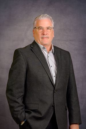 Professional headshot of a middle-aged man with gray hair, glasses, and a dark blazer over a patterned shirt.